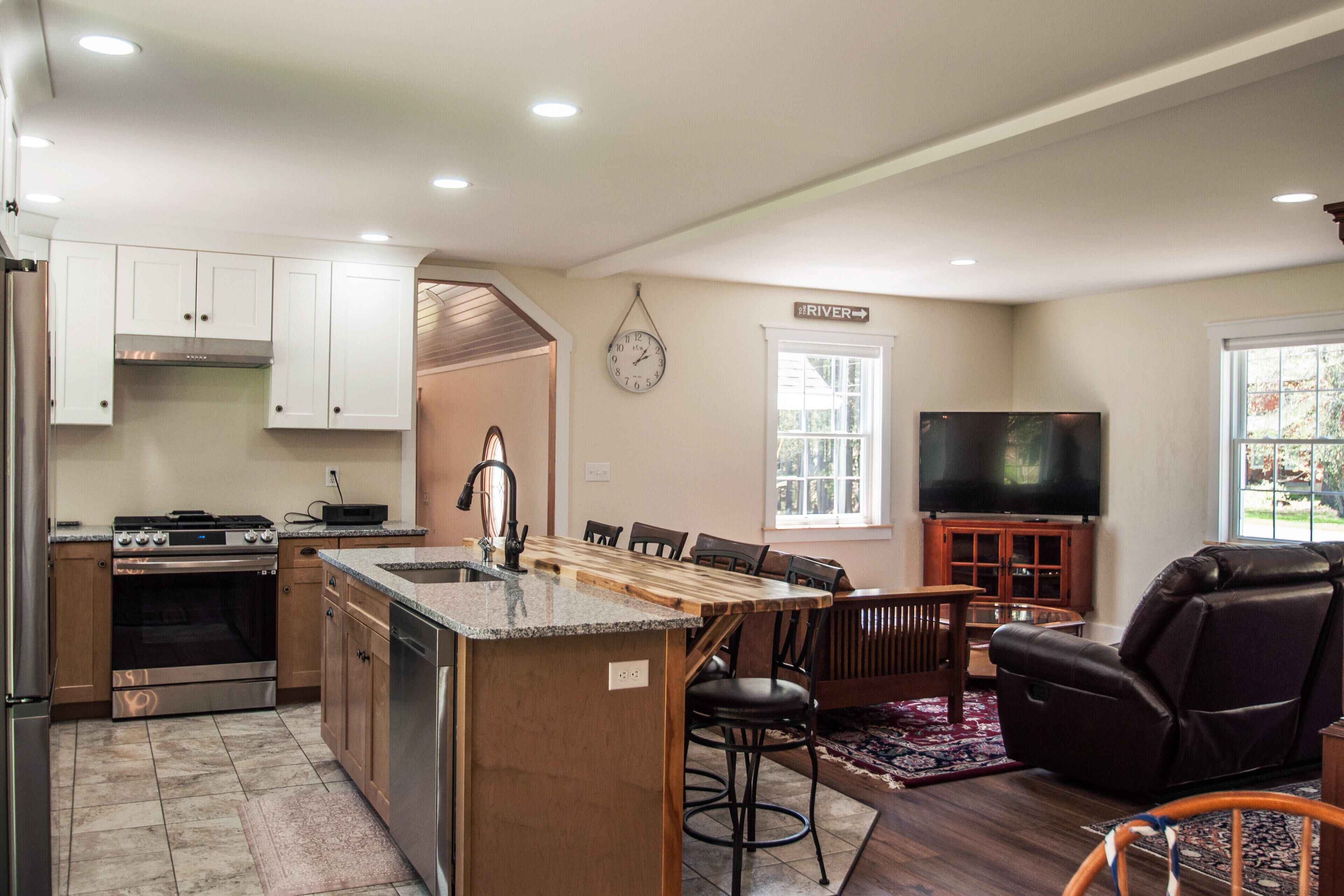 A modern kitchen with a central island, a dining area with chairs, a flat-screen TV, and a large window.