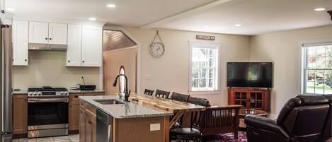 A modern kitchen with a central island, a dining area with chairs, a flat-screen TV, and a large window.