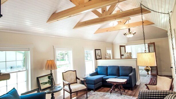 A spacious living room with a high ceiling, wooden beams, a blue sofa, a patterned armchair, and a chandelier.