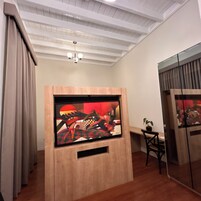 Una habitación con un mueble bajo de televisión de madera, techo con vigas a la vista, una araña, un escritorio con una silla y una planta en maceta.