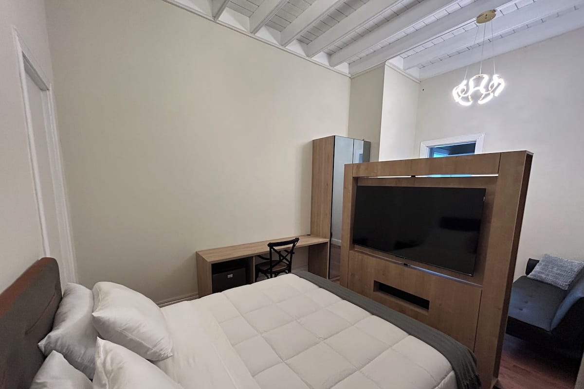 A bedroom with a bed, a TV mounted on a wooden cabinet, a desk with a chair, and a neon sign on the wall.