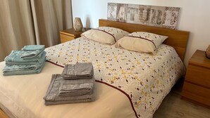 Una cama bien hecha con ropa de cama estampada, dos almohadas y toallas dobladas sobre una mesita de noche de madera.