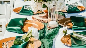 Una mesa dispuesta con servilletas verdes, platos anaranjados y un centro de mesa con flores blancas en un florero de vidrio.
