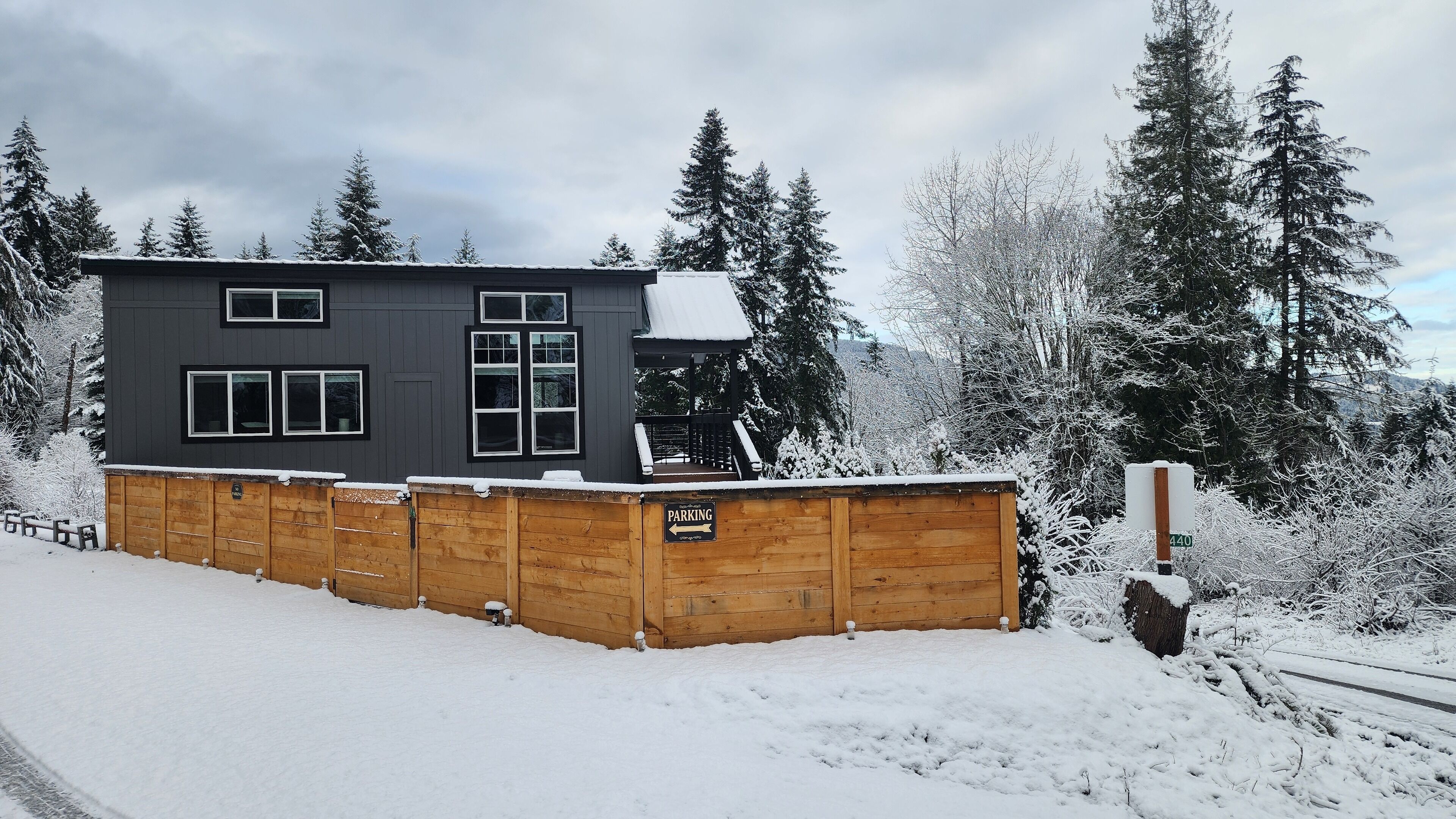 A modern two-story building with a dark exterior, surrounded by snow-covered trees and a wooden fence.