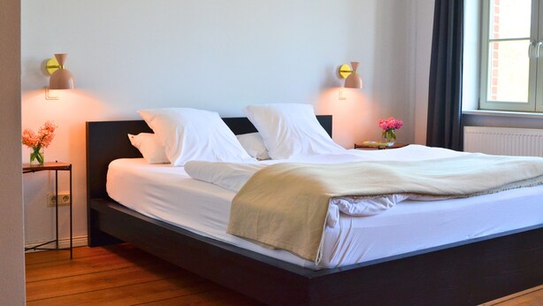 Een net opgemaakt bed met witte lakens en een beige deken, met aan weerszijden nachtkastjes met lampen en bloemen.'