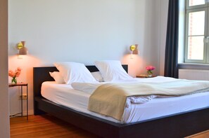 A neatly made bed with white linens and a beige throw blanket, flanked by two bedside tables with lamps and vases of flowers. - 3-bed room in the Gasthaus am Finowkanal (former school from 1875). (Wandlitz)
