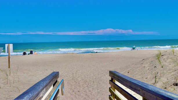 A beach with a wooden walkway leading to the water, a clear sky, and a distant view of the ocean.