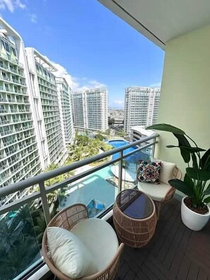 A balcony with wicker furniture, a potted plant, and a view of high-rise buildings and a pool. - Azure Create Greate Moments by Don (Paranaque)