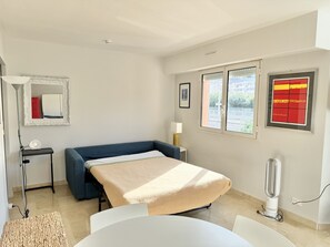 Una camera d'albergo compatta con un letto singolo, una piccola scrivania, una sedia, una lampada da terra e uno specchio fissato al muro, oltre a una finestra con vista.