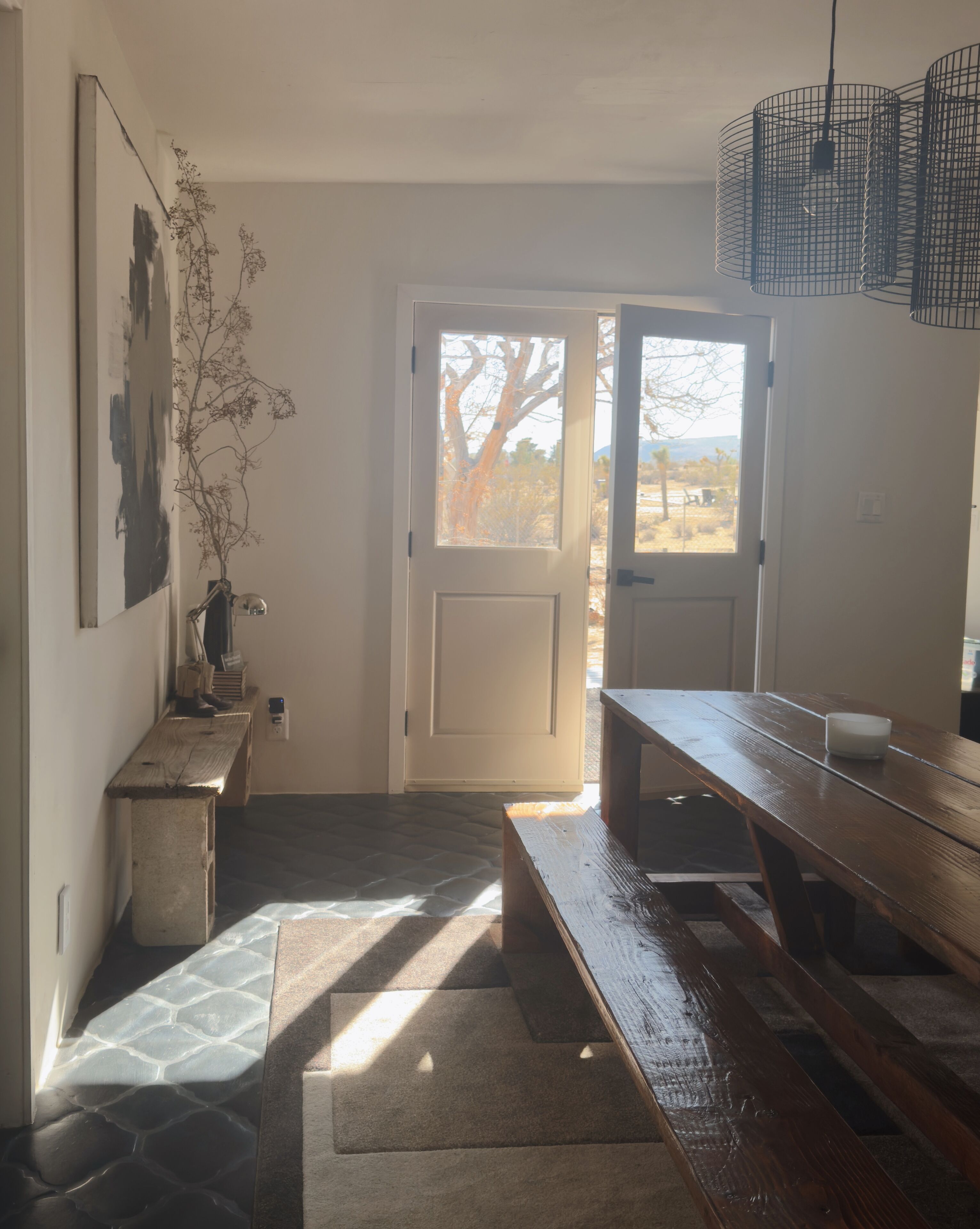 A room with a wooden bench, a table, a vase with branches, and a door leading outside.