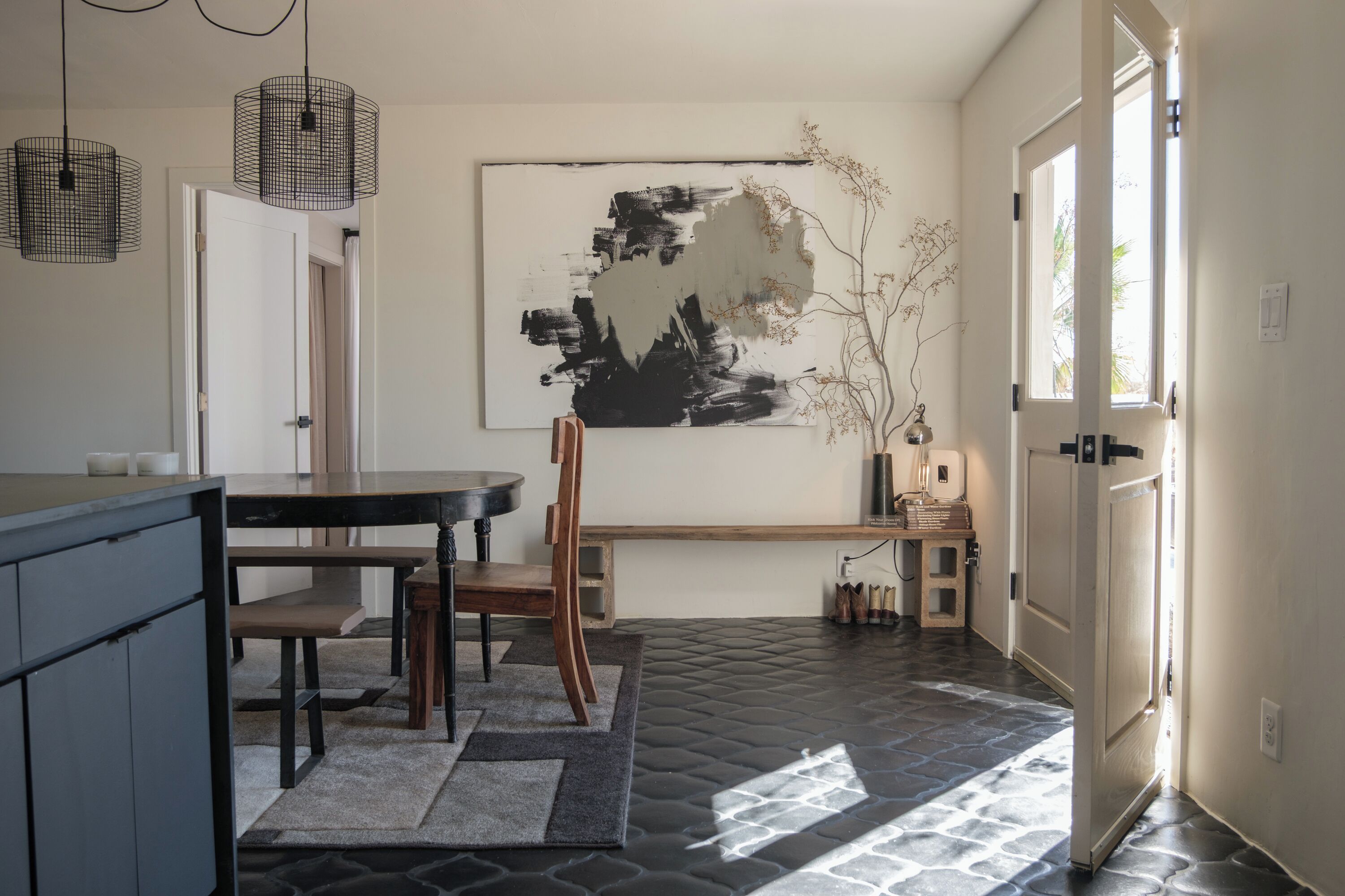 A modern kitchen with a dining area, featuring a black and white abstract painting, a wooden bench, and a stone-patterned floor.