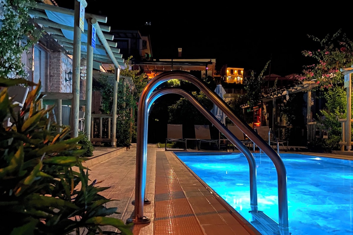 Ein Schwimmbeckenbereich bei Nacht mit einer Metallleiter, die ins Wasser führt, umgeben von Grünpflanzen und Gebäuden.