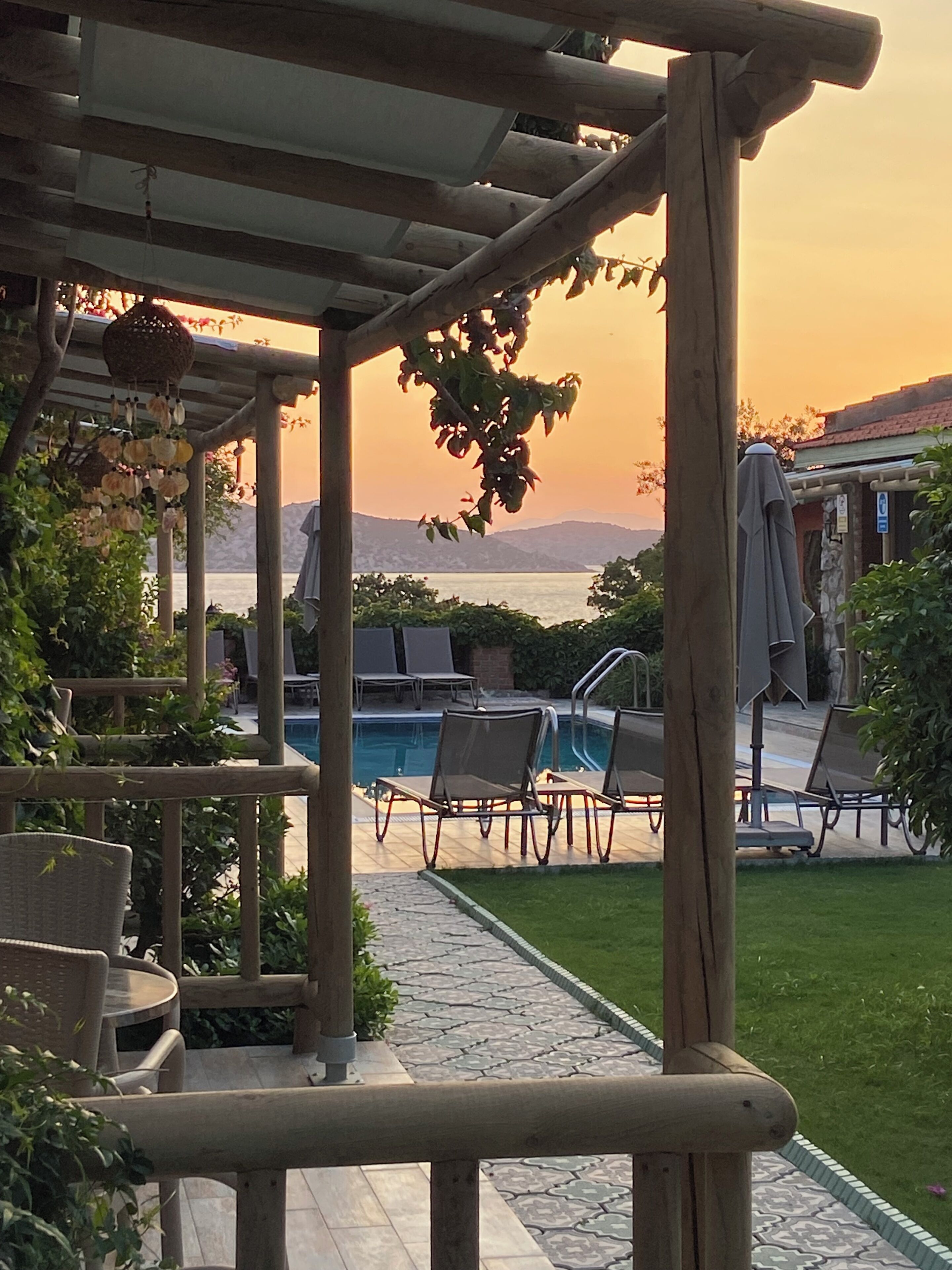 A poolside area with lounge chairs, a pergola, and a view of the sea at sunset.