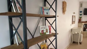 Una estantería con libros y adornos, una silla blanca y una pequeña mesa en una habitación con suelo de baldosas y pared blanca.