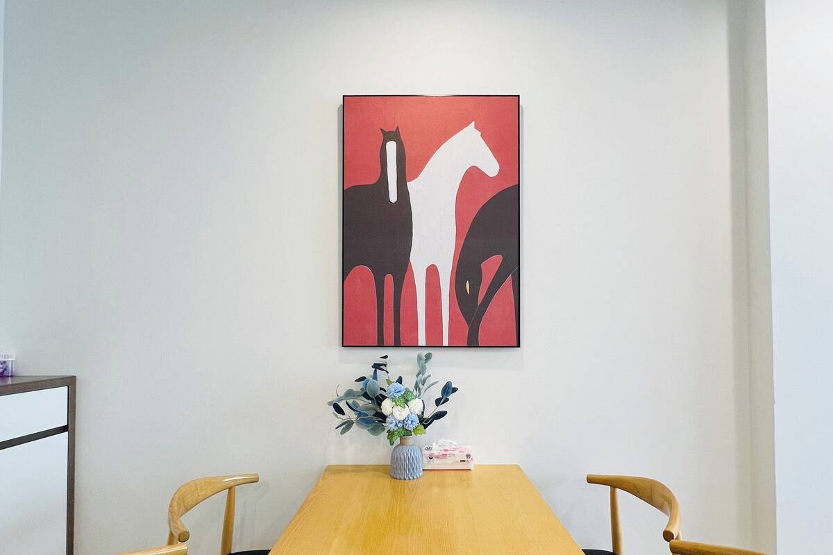 Een eetgedeelte met een houten tafel, zwarte stoelen met kussens, een vaas met bloemen en een kunstwerk met rode en zwarte kleuren aan de muur.