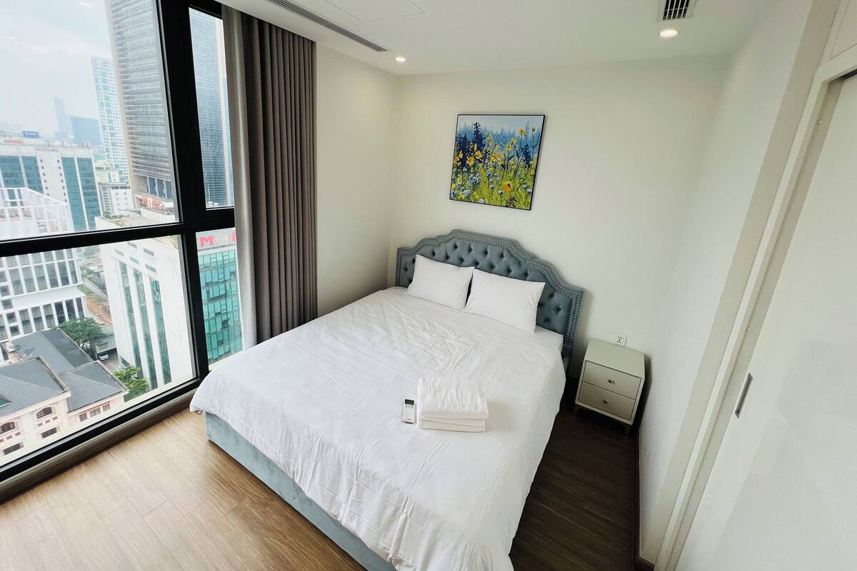Een moderne hotelkamer met een groot bed, een nachtkastje, een schilderij en uitzicht op de stad door grote ramen.