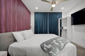 A hotel room with a bed, a ceiling fan, a TV mounted on the wall, and a window with curtains.