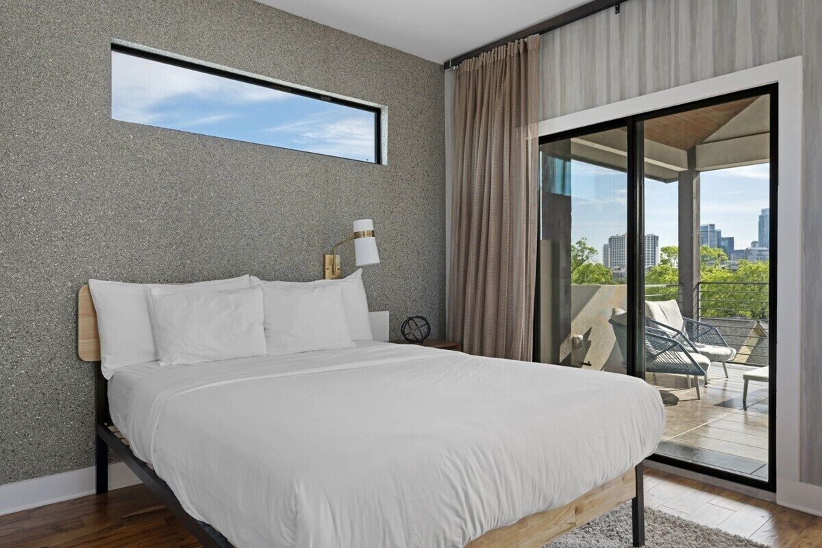 A modern bedroom with a large bed, a nightstand, a lamp, and a view of the city through a glass door.