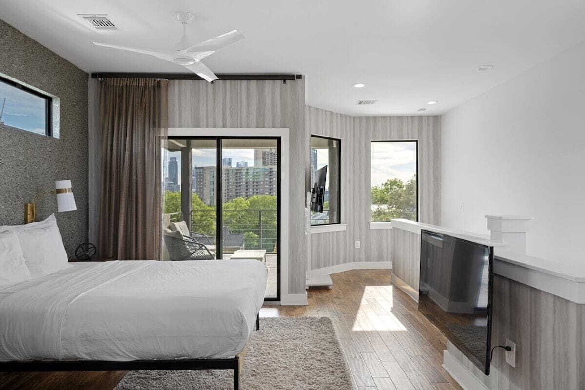 A modern bedroom with a large bed, a ceiling fan, and a view of a cityscape through a sliding door.