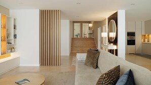 A modern living room with a sofa, a coffee table, and a bookshelf. There is a wooden slat partition and a kitchen area in the background.