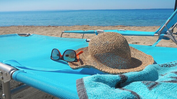 Ein Strandstuhl mit Strohhut und Sonnenbrille auf einer blauen Unterlage, mit Blick aufs Meer.