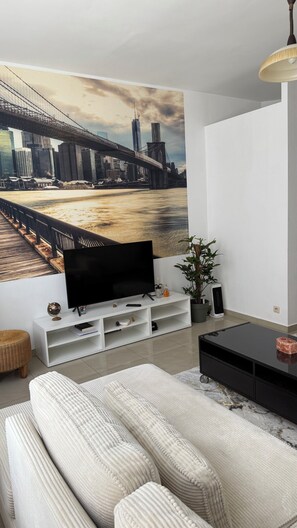 Un salon moderne avec une grande fresque représentant un paysage urbain, un téléviseur à écran plat, un canapé blanc et une table basse noire.