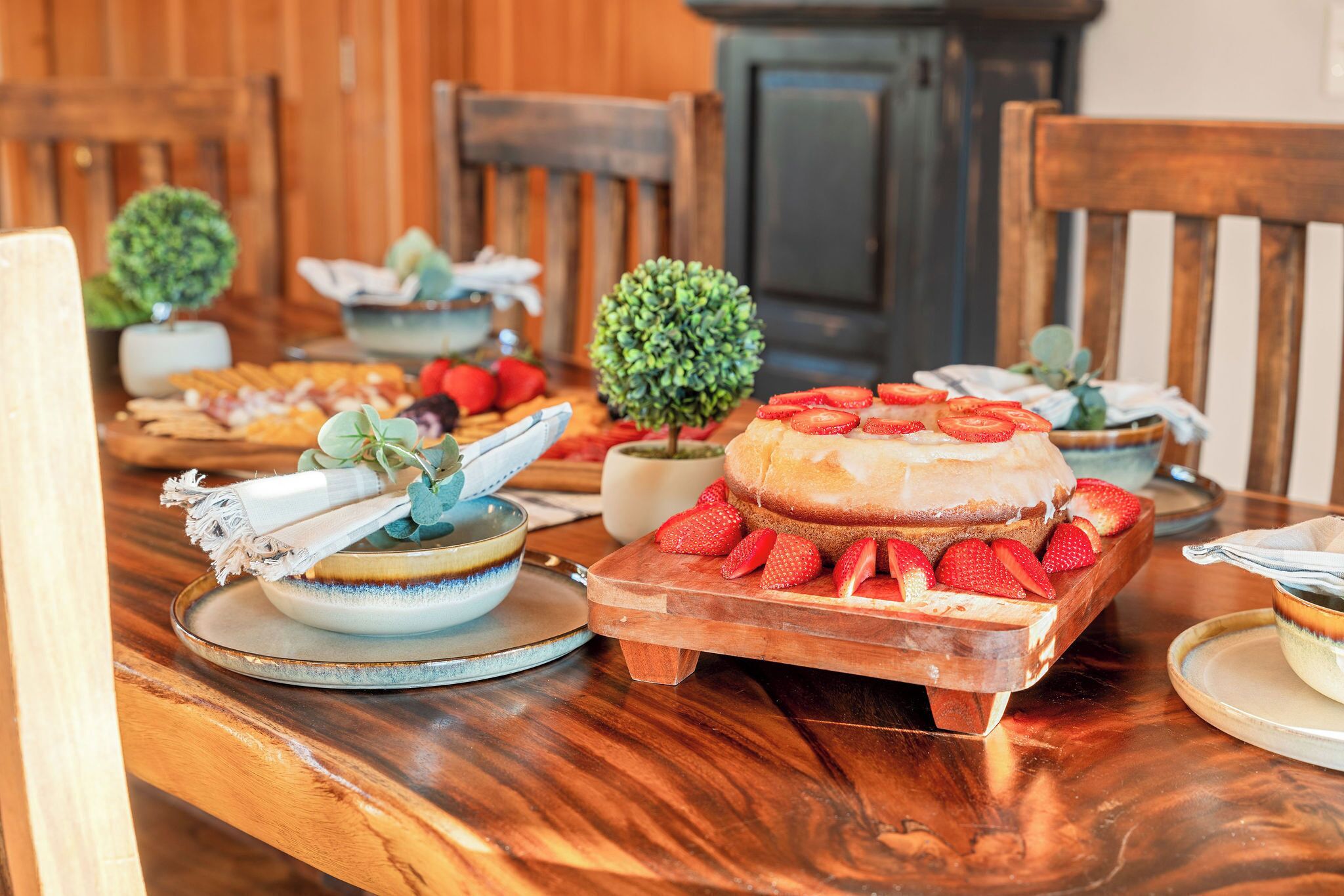 Een houten eettafel met verschillende soorten eten, waaronder een taart met aardbeien, kommen en een kleine plant in een pot.