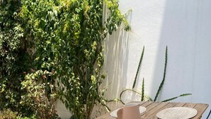 A wooden table with woven placemats and a pitcher, set outdoors with a white wall and greenery in the background.