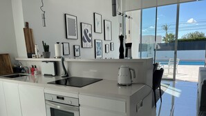 A modern kitchen with a built-in oven, a kettle, and a coffee maker. There are framed pictures on the wall and a large window offering a view of the outside. - Elegant functional villa with saltwater pool (Torrevieja)