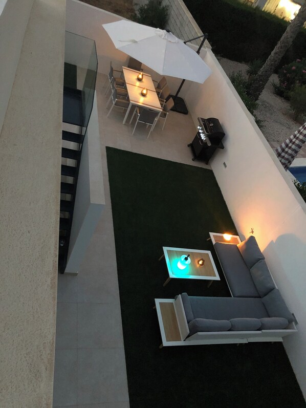 An outdoor patio area with a glass balustrade, a dining table with chairs, a sofa, and a small table with a light.
