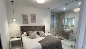 A modern bedroom with a large bed, bedside tables, a nightstand, a bedside lamp, a bedside plant, and a bathroom visible through a glass door. - Elegant functional villa with saltwater pool (Torrevieja)