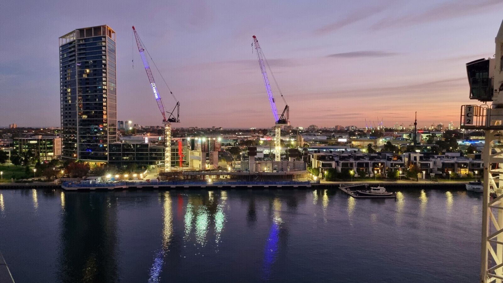 A waterfront cityscape with modern buildings, cranes, and a marina.