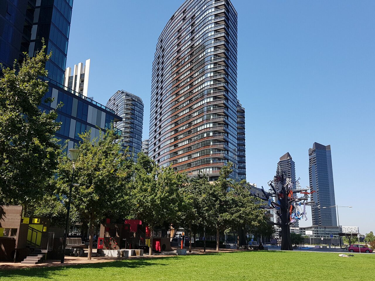 A modern urban park with a grassy area, trees, and tall buildings in the background.