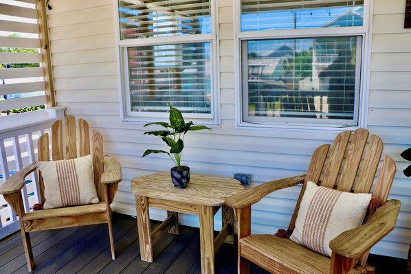A wooden deck with two chairs and a small table, a potted plant, and a window with blinds. - Surf & Sun Haven: Steps to Sea-Clusion (Kure Beach)