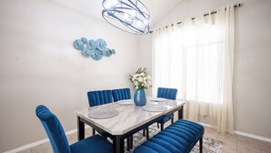 A dining area with a marble table, blue upholstered chairs, a blue bench, a vase with white flowers, a blue chandelier, and a window with sheer curtains. - Chic Vegas House w/Game Room + BBQ (North Las Vegas)