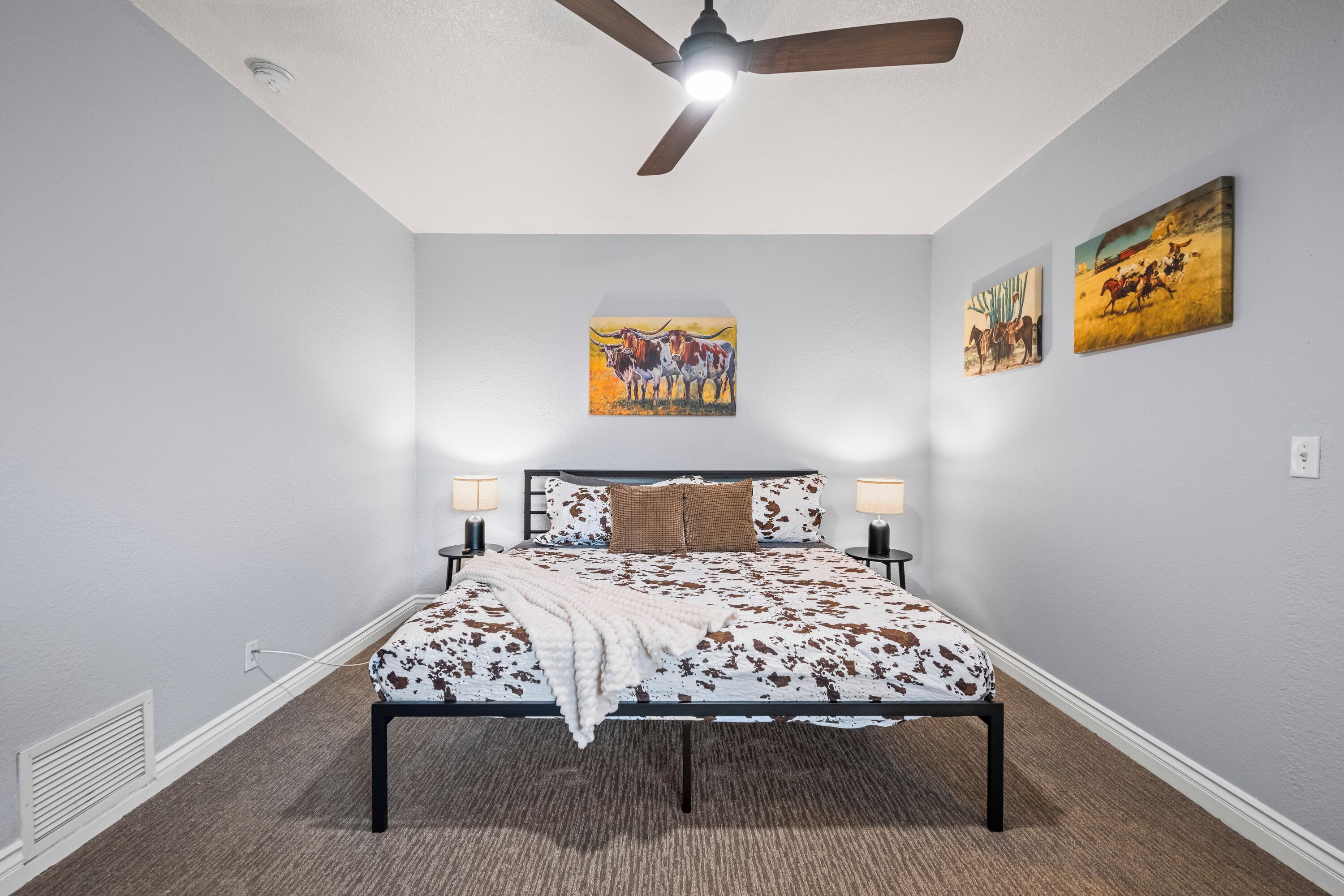 A bedroom with a bed, ceiling fan, wall-mounted artwork, bedside lamps, and a carpeted floor.