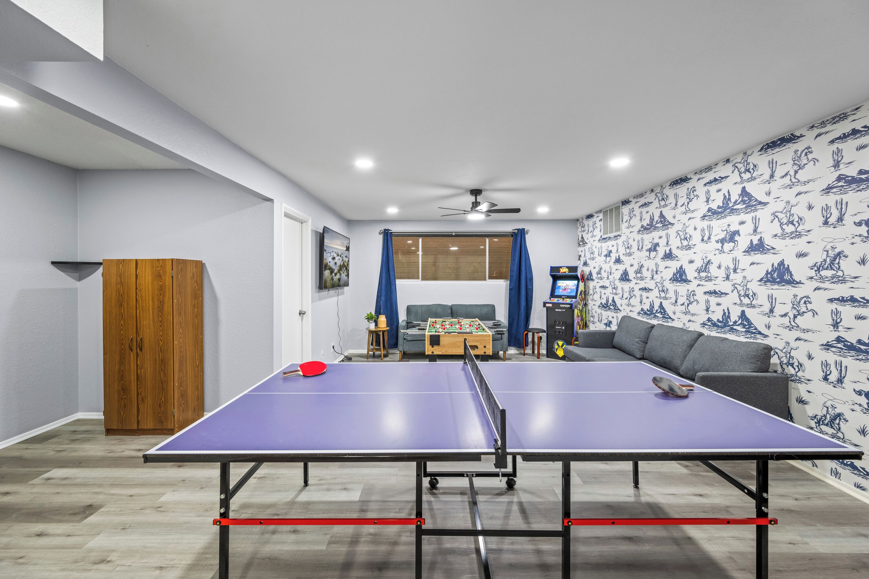 A room with a ping pong table, a wooden cabinet, a sofa, and a wall with a desert-themed wallpaper.