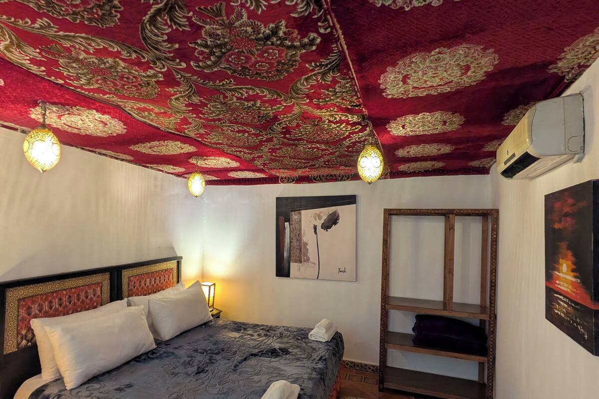 A room with a bed, a wall-mounted air conditioner, a framed picture, and a patterned carpet.
