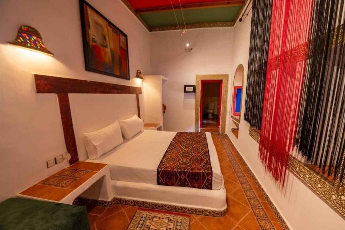 A hotel room with a bed, a nightstand, a wall-mounted lamp, a patterned rug, and a decorative wall hanging.