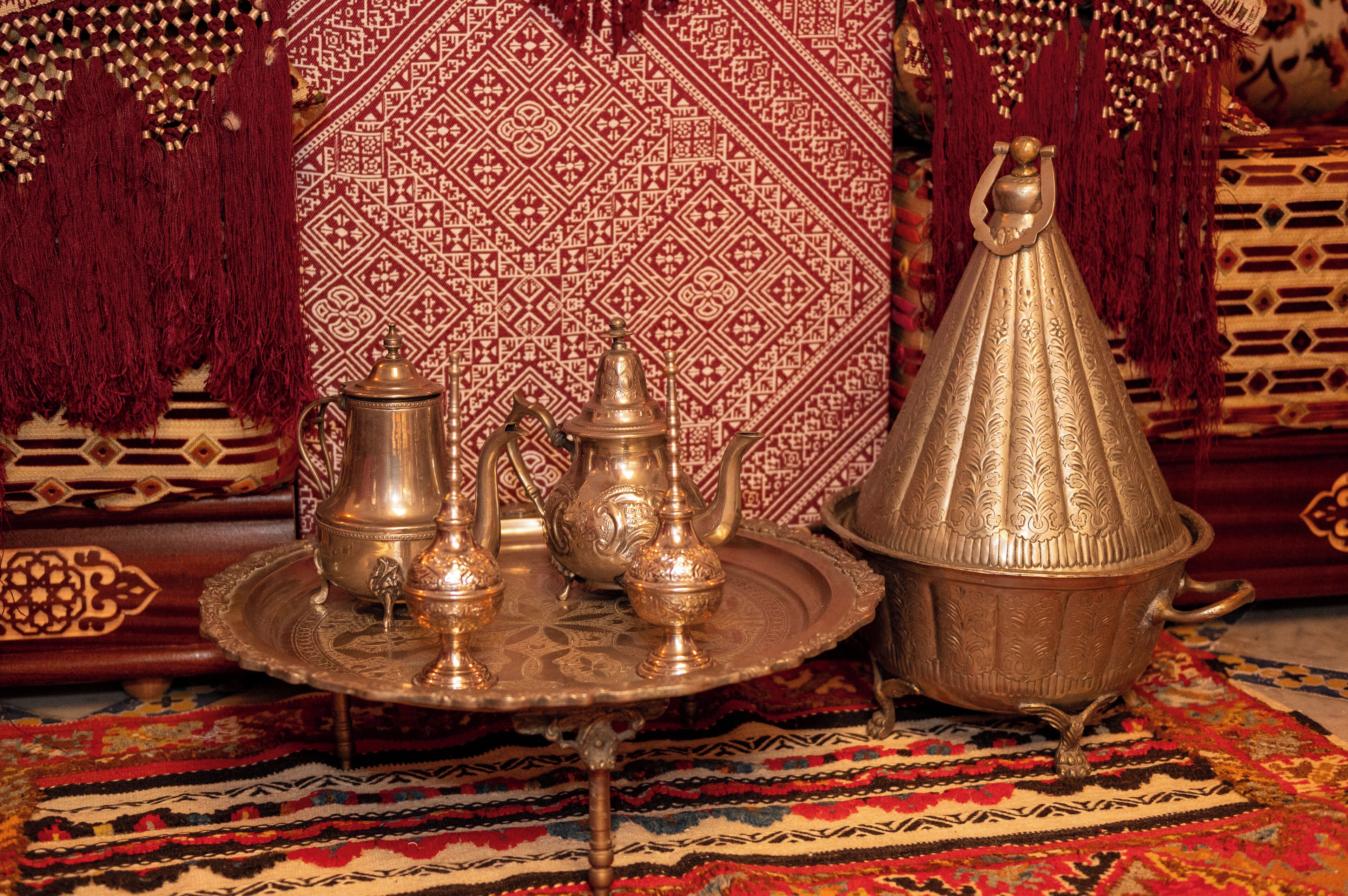 Um ambiente tradicional marroquino com um bule de cobre, chaleira e açucareiro em um tabuleiro, um tagine de cobre grande e ornamentado, e tecidos com padrões vibrantes.