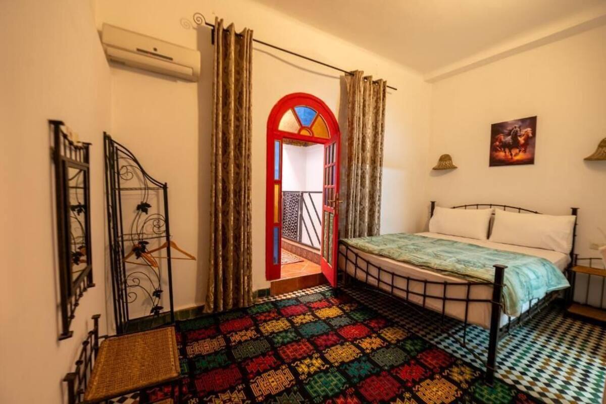 A bedroom with a bed, a colorful rug, a red-framed door, and a painting on the wall.