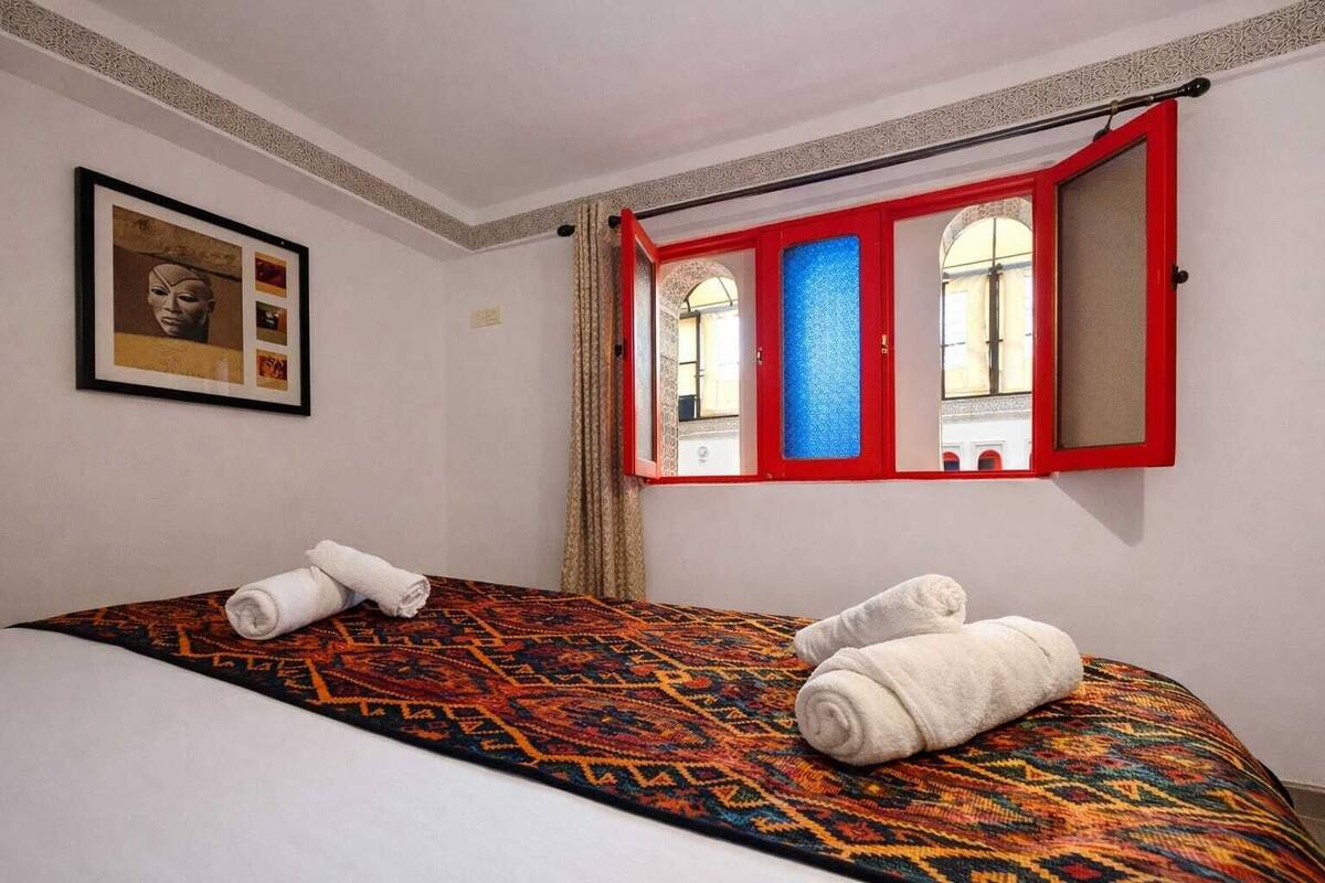 A hotel room with a bed, two rolled towels, a framed picture on the wall, and a window with red frames.