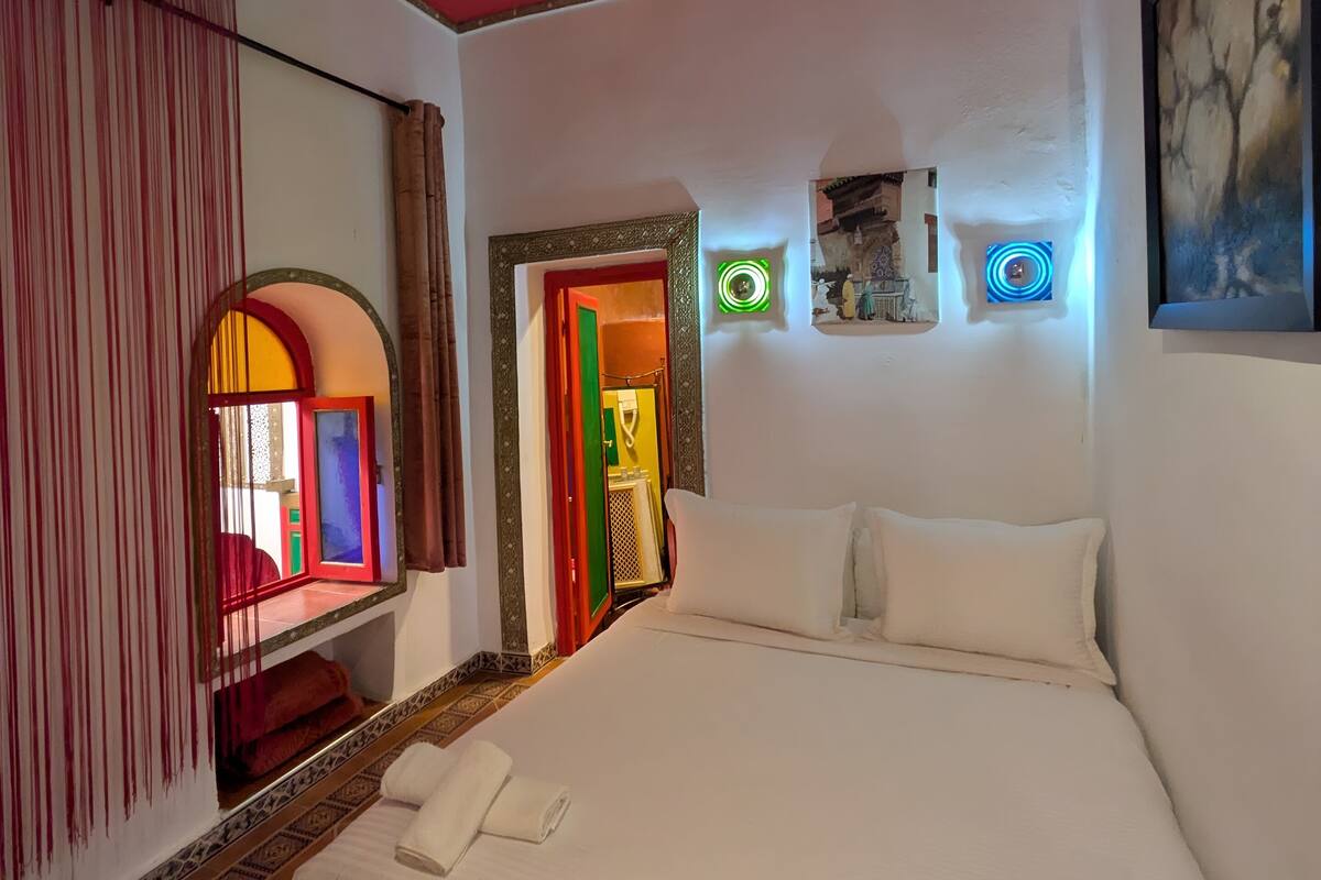 A hotel room with a large bed, a mirror, a painting, and a view of a colorful interior through an open door.