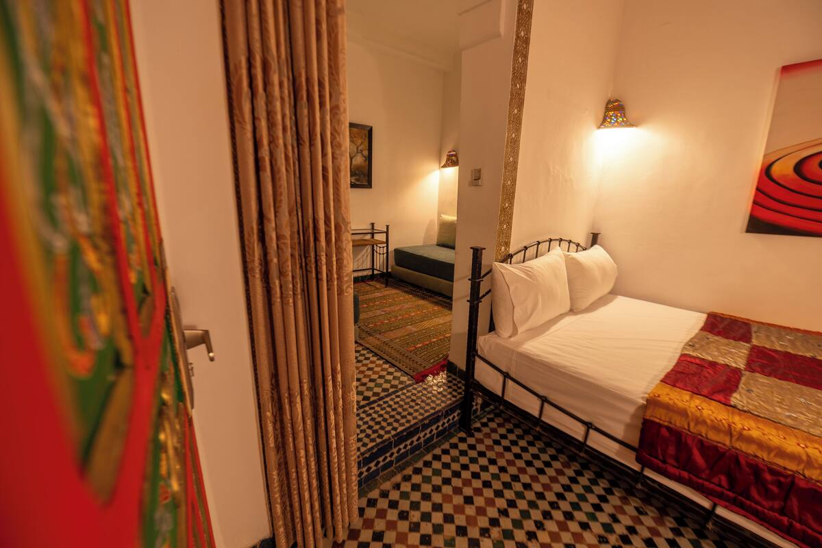 A bedroom with a checkered floor, a bed with white linens, a patterned curtain, and a wall-mounted light fixture.