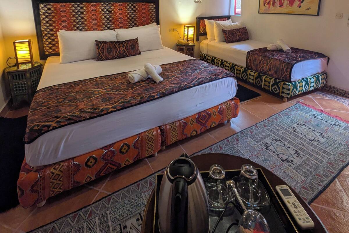 A hotel room with a large bed, a smaller bed, a sofa, a coffee table with a kettle, glasses, and a remote control.