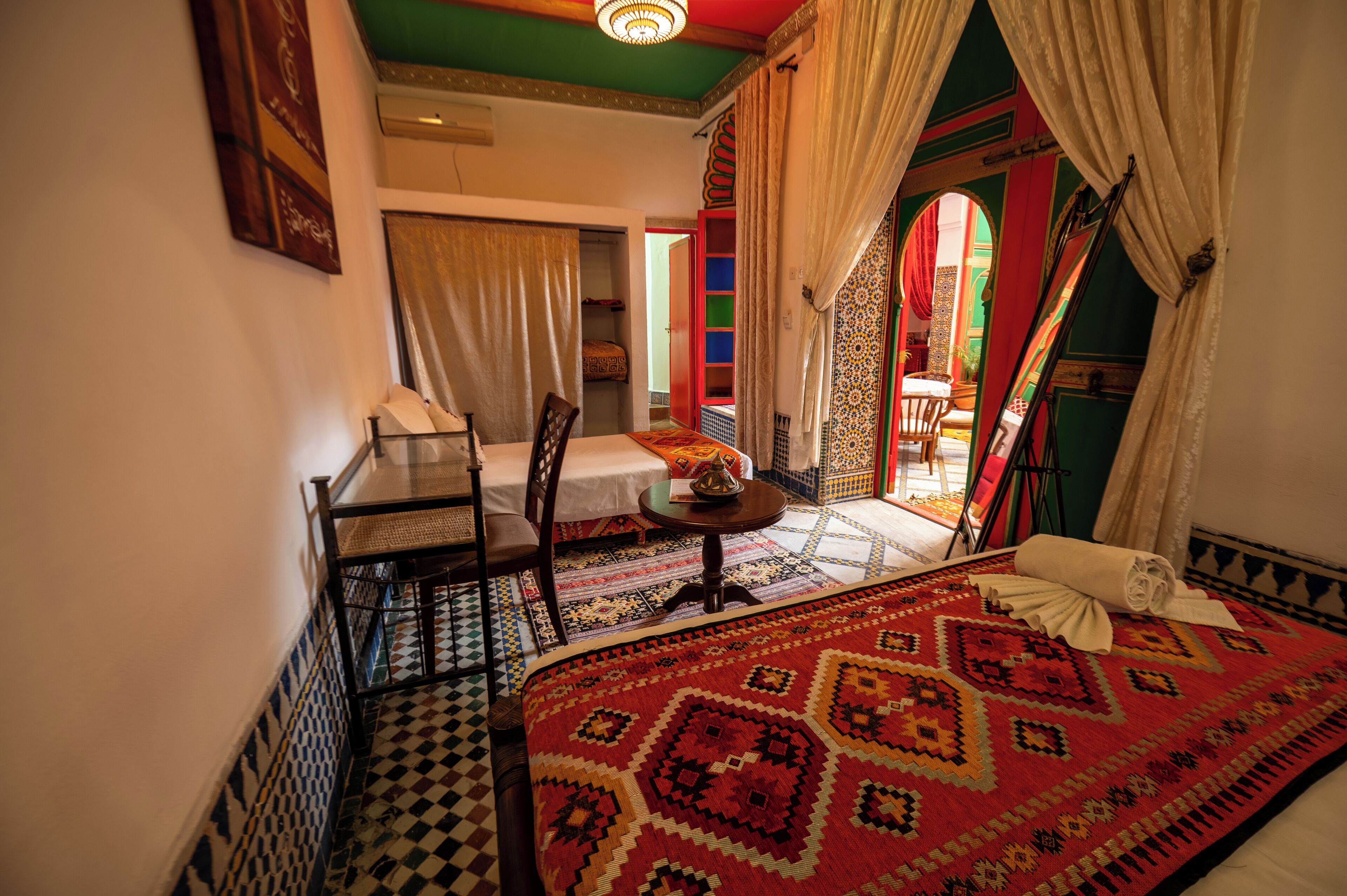 A room with a bed, a table, and a chair. There is a window with curtains, a door with a colorful frame, and a patterned rug on the floor.