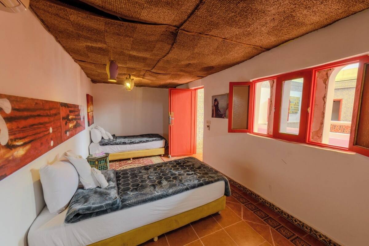 A room with two beds, a thatched ceiling, and a window with red frames.