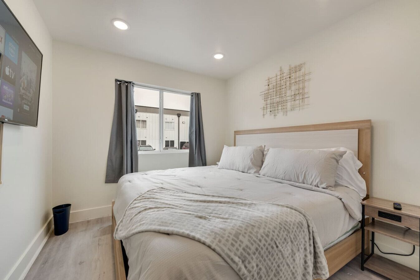 Un dormitorio con cama, mesita de noche, ventana con cortinas y una obra de arte en la pared.