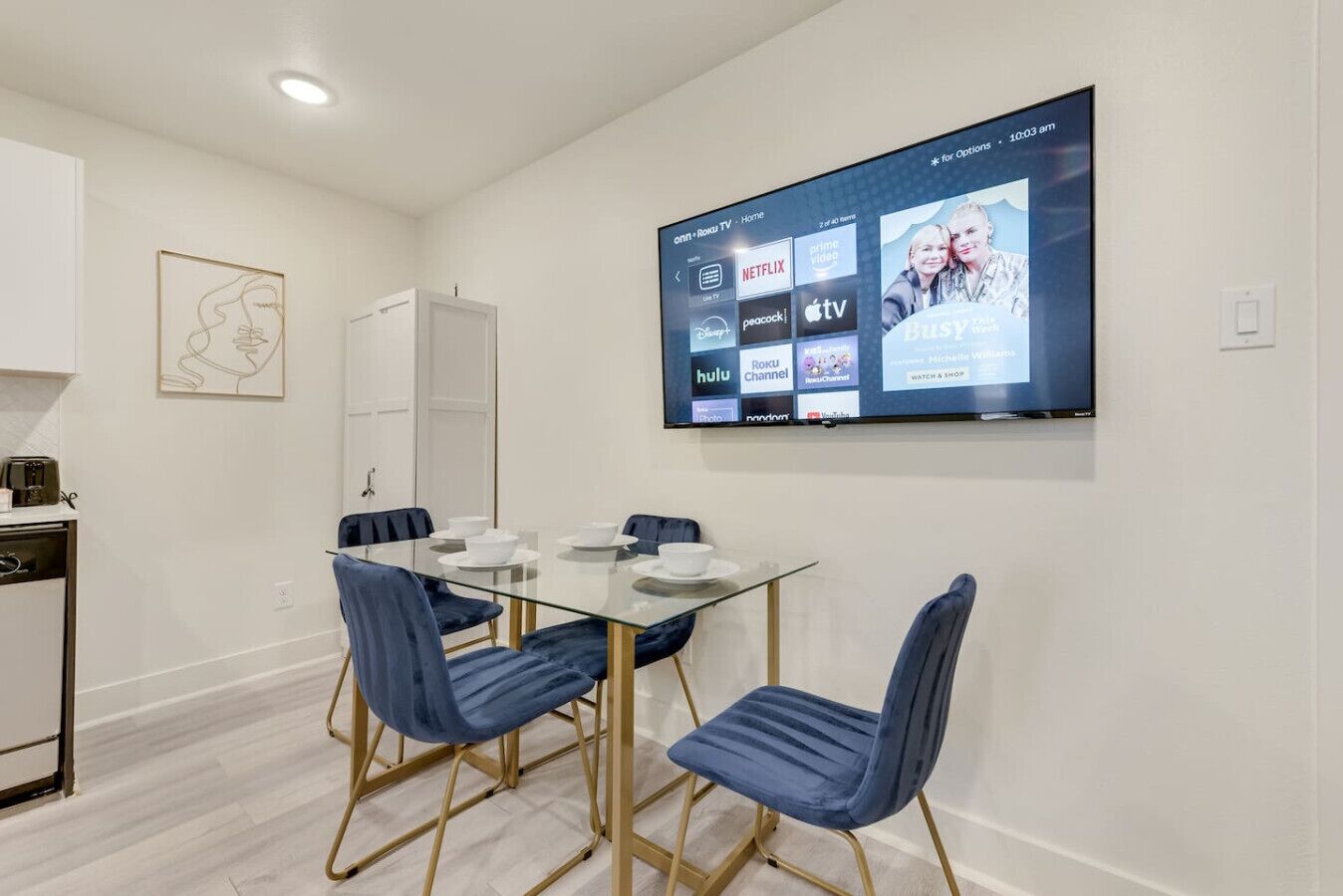 Un comedor moderno con una mesa de vidrio, sillas tapizadas en azul y un televisor de pantalla plana mostrando servicios de transmisión.