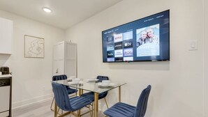 A modern dining area with a glass table, blue upholstered chairs, and a flat-screen TV displaying streaming services. - Stylish City Getaway | Near Deep Ellum & Downtown (Dallas)